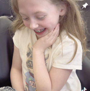 A girl with braces, wearing a light-colored shirt, smiles and looks down with her hand on her cheek.