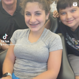 Two children smiling while sitting, with a blurred adult in the background.