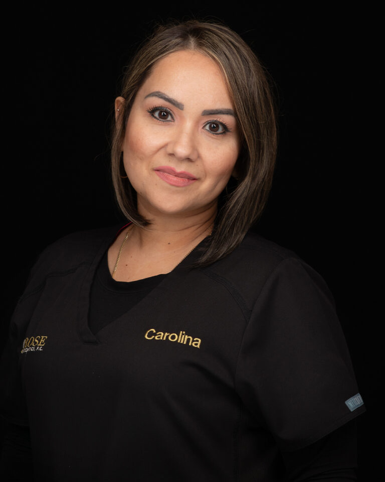 Carolina, a woman with shoulder-length hair wearing black scrubs, stands against a black background.