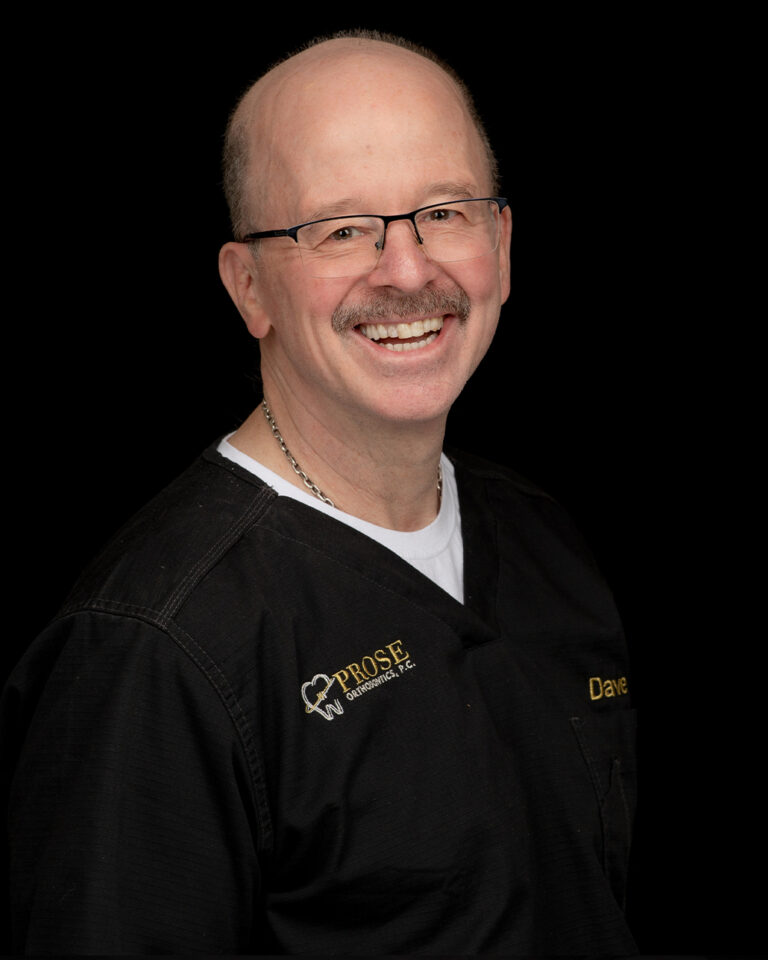 Dave, a man wearing glasses, black scrubs, and a white undershirt smiles against a black background.