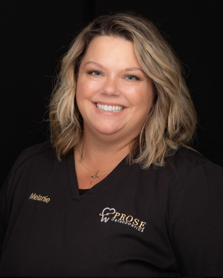 Melanie, a woman with shoulder-length hair in black scrubs, stands against a black background.