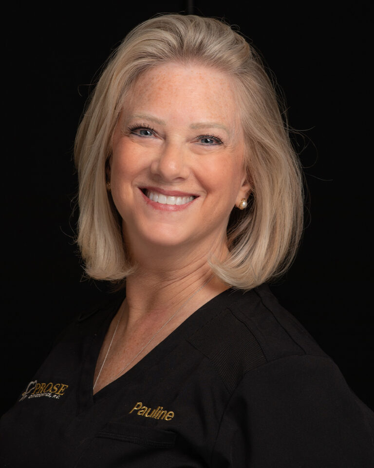 Pauline, a woman with short blonde hair in black scrubs, stands against a black background.