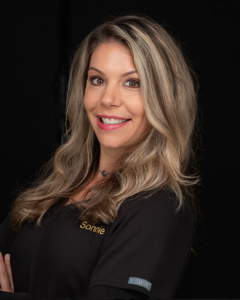 Sonnie, a woman with long blonde hair in black scrubs, stands against a black background.