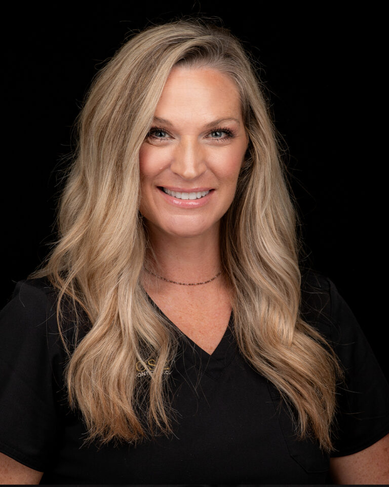 Tae, a woman with long blonde hair in black scrubs, stands against a black background.