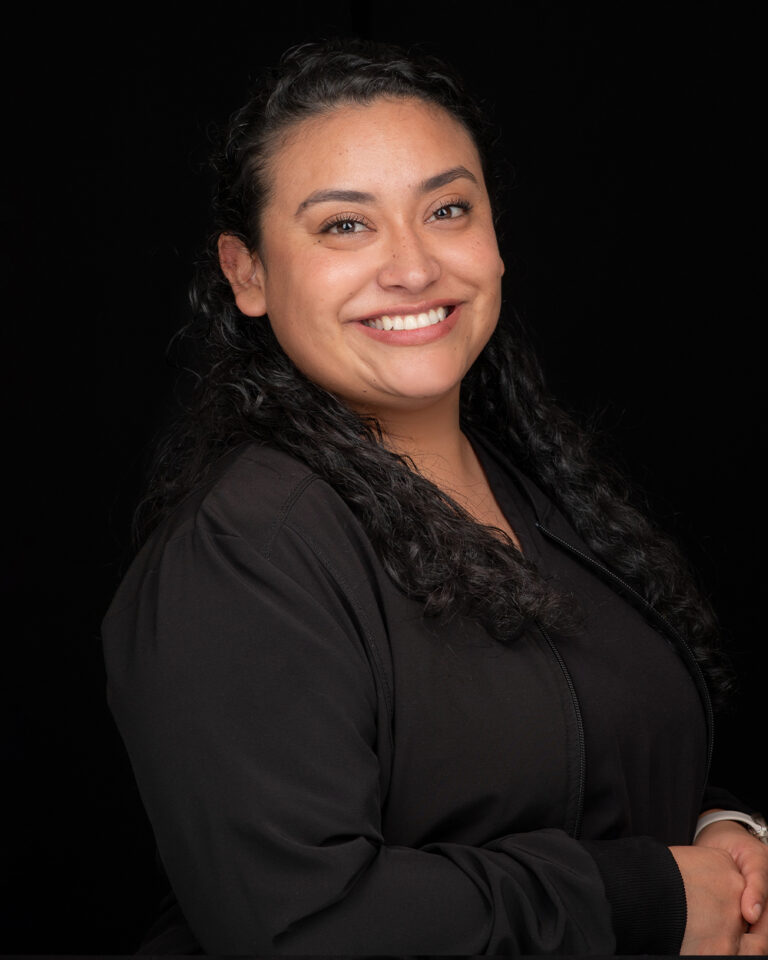 Yessi, a woman with curly black hair in black scrubs, stands against a black background.