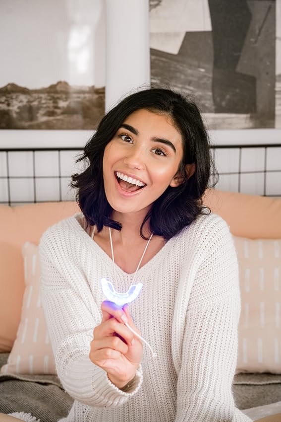 Young woman holding her GLO Whitening LED Mouthpiece while smiling on her bed
