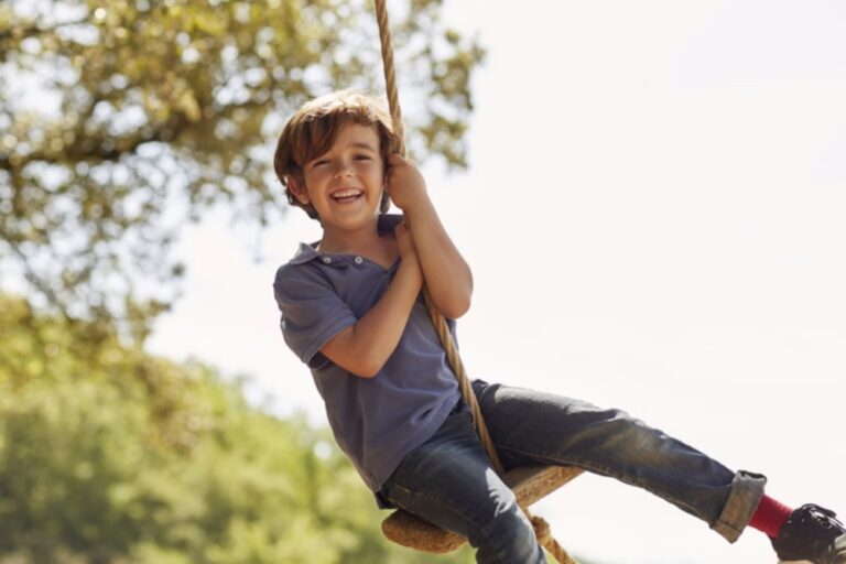 Portrait of cheerful boy playing on swing. Happy male is wearing casuals. He is enjoying against clear sky.