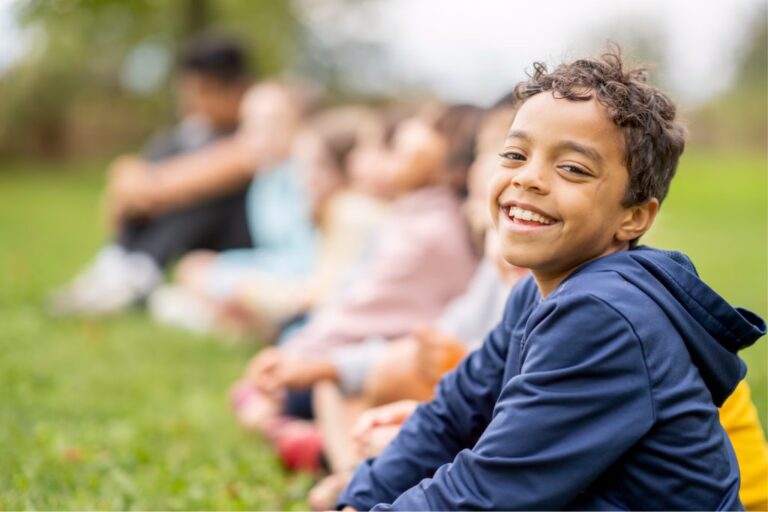 A tan boy sitting in the grass, looking at the camera smiling in a blue hoodie.