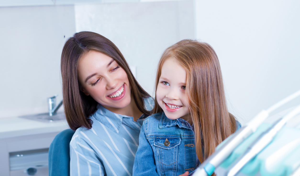 Little cute smiling girl is sitting in dental chair with mother in clinic, office. Doctor is preparing for examination of child teeth, talking with patient. Visiting dentist with children.