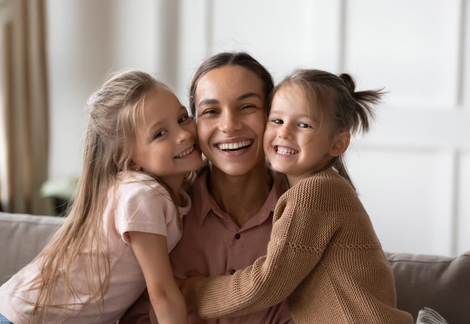Beautiful female family young smiling single mother and cute little children daughters embracing looking at camera, funny small kids girls hugging happy mum bonding sit on sofa together, portrait